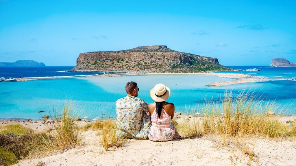A couple sitting on a hill looking at an island direct infront of them sourounded by cristalclear blue waters.