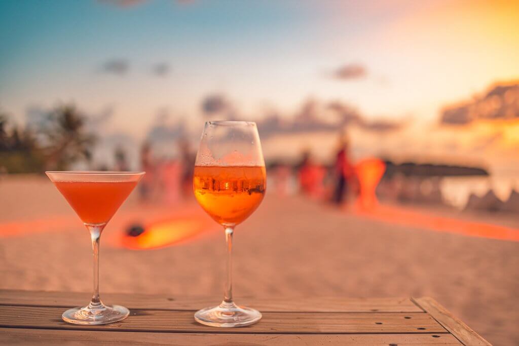 Two cocktail glasses filled with a red drink with the sunset on a beach in the background.