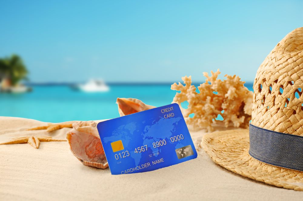 A travel credit card lying in the sand on the beach next to coral and blue sea in the back.