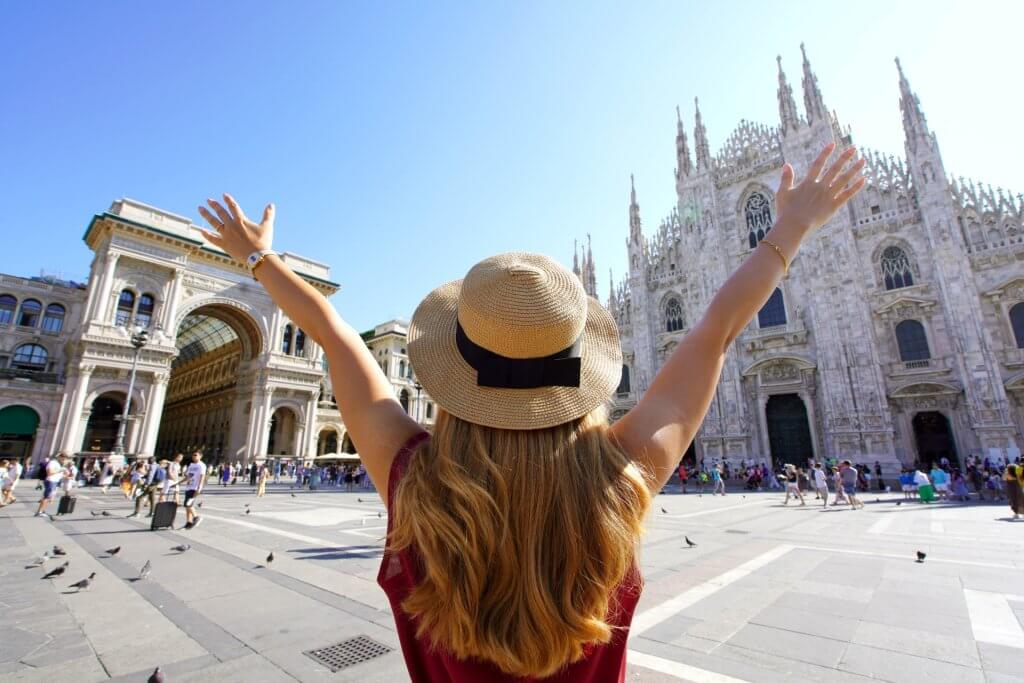 Young woman with a summer hat opens both arms in the air infront of church in Milan.
