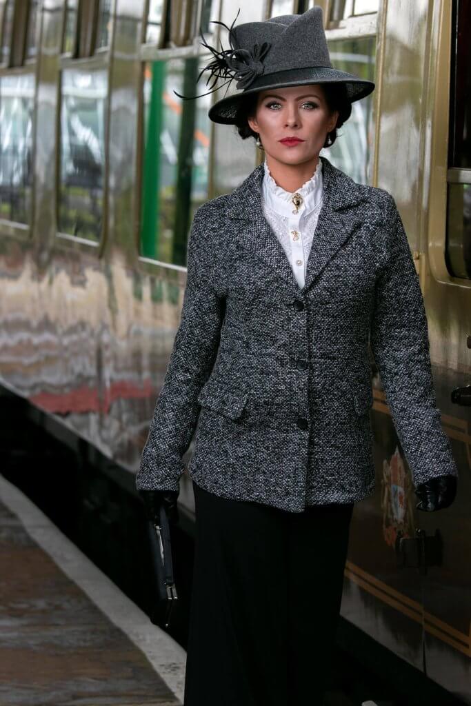 An elegant woman wears a tweed blazer, black skirt and a black hat.