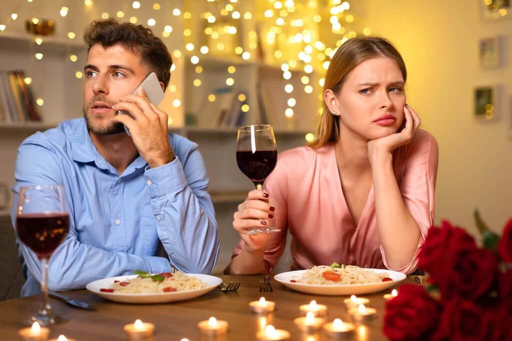 A couple is sitting at the table. He is on the phone and she has an annoyed look on her face.