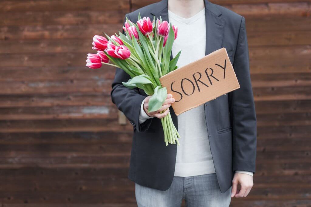 A man holds a bouquet of flowers and a sign that says Sorry.
