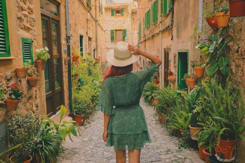 A young woman with a grren dress and a white hat walking in a street full of flowers.