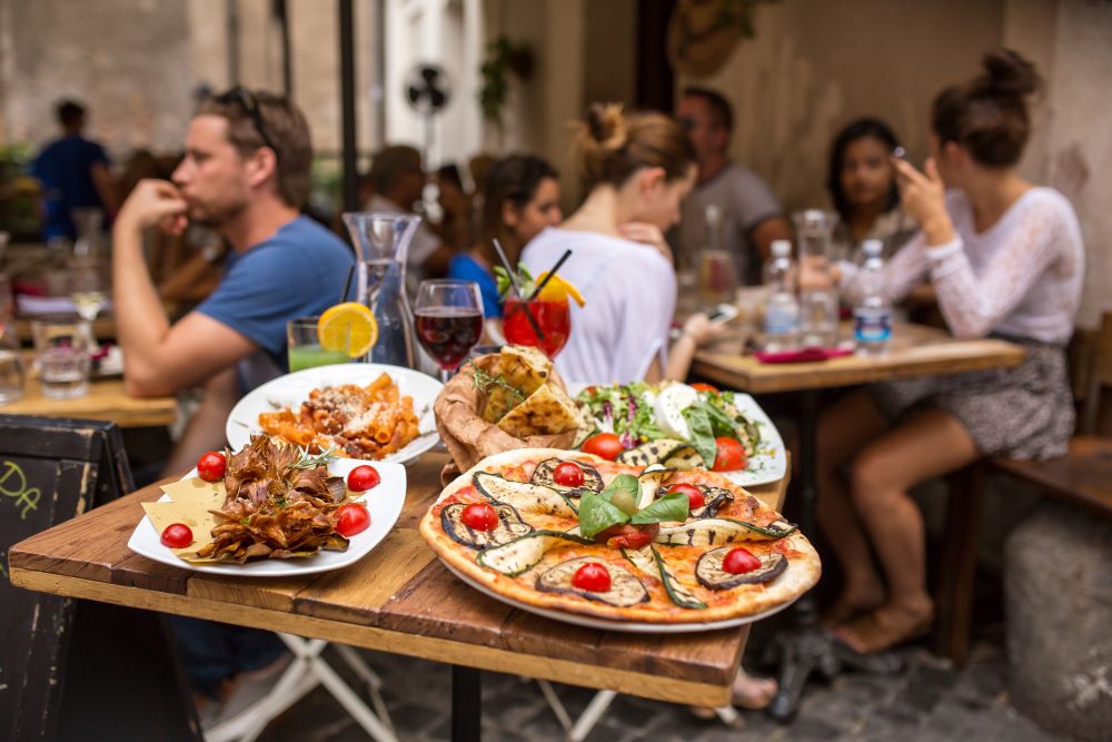 Outdoor terrace with people and a table in the foreground with pizza, pasta and drinks.