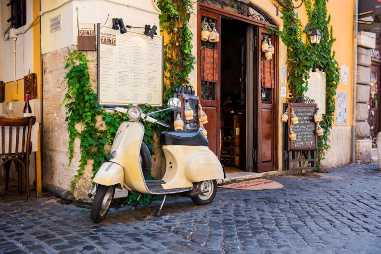 A beige Vespa scooter stands in front of a flower-covered house in Rome.
