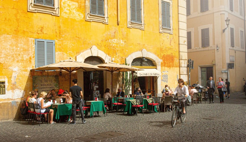 Outdoor terrace infront of a big yellow house with people sitting outsie having lunch.