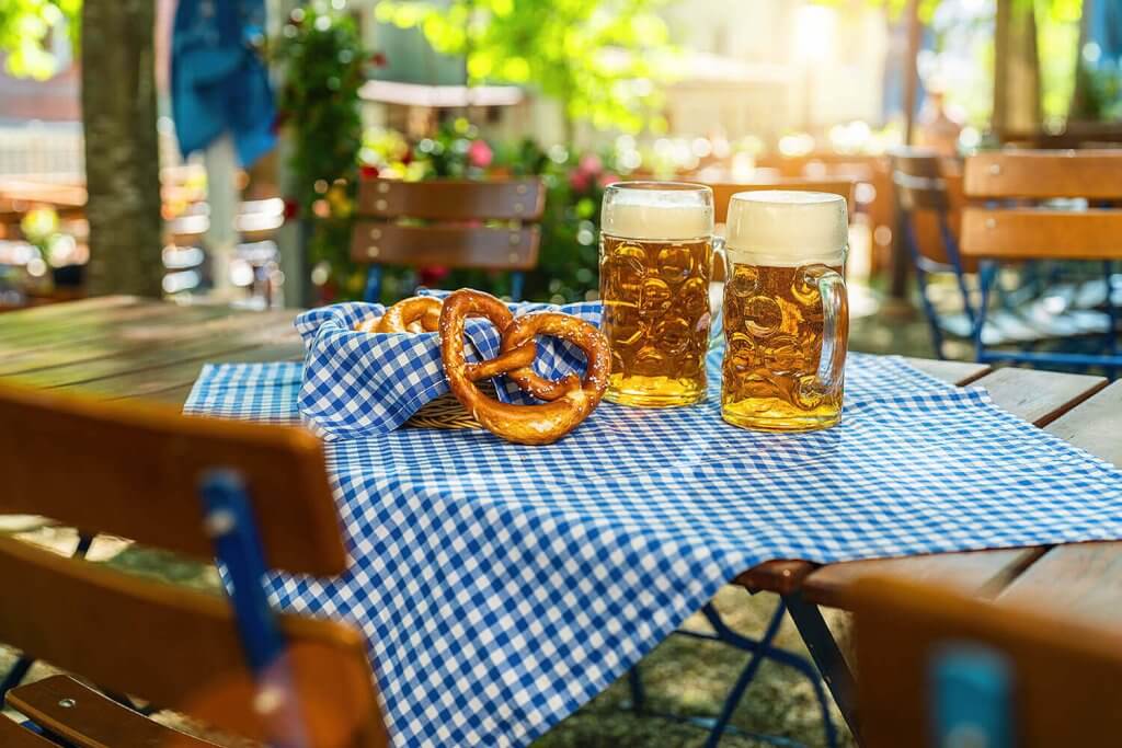 A table in a beer garden with two pints of beer and a pretzel.