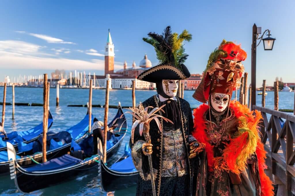 Two traditionally costumed people with masks in Venice with gondolas in the background.