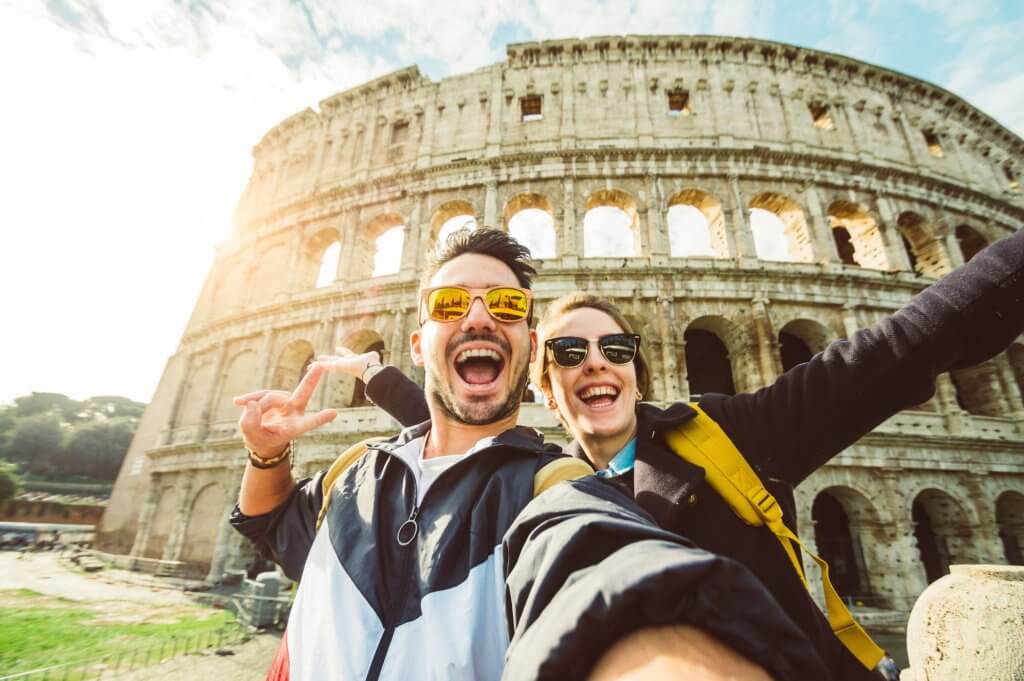 A young couple takes a selfie right in front of the Colloseum in Rome.